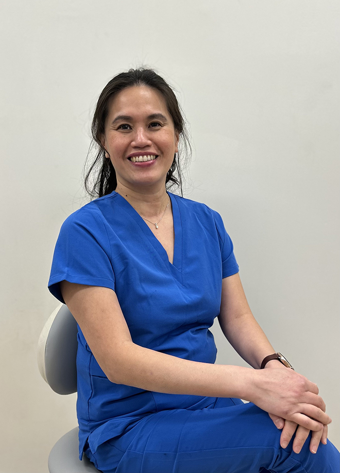A smiling female nurse seated on a stool in front of a white wall.