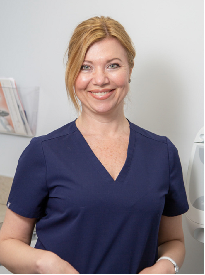 A woman wearing a navy blue scrub top, standing with a slight smile, in front of a white wall with a shelf holding items, and a window behind her.