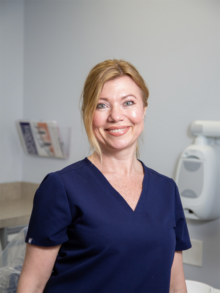 The image shows a woman wearing scrubs, standing in an office environment with medical equipment visible behind her.