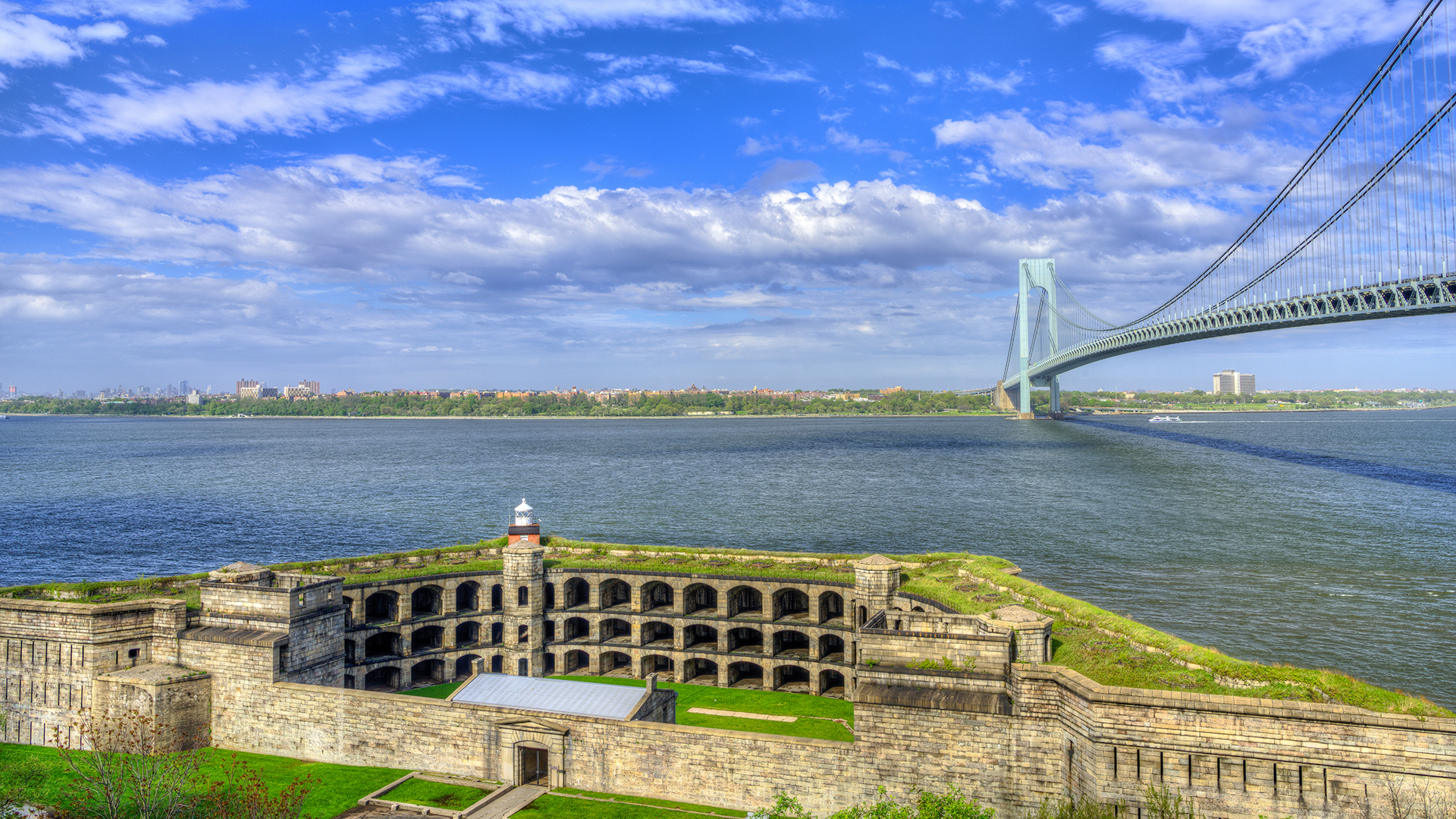 The image features a large, imposing brick building situated on an island with a bridge spanning across water in the background under a clear blue sky.