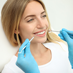 The image shows a woman sitting in a dental chair with a smiling expression while receiving dental care, with a dental hygienist performing a cleaning procedure visible through her mouth.
