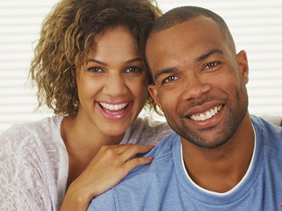 The image shows a man and a woman smiling at the camera  the man is wearing a short beard and has his arm around the woman, who is laughing. They appear to be posing for a portrait-style photograph with a warm and happy expression.